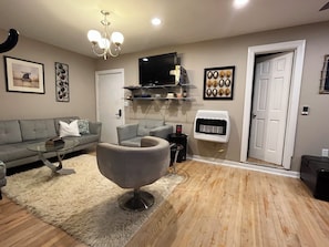 A living room with a grey sofa, a glass coffee table, a modern armchair, a white radiator, and a flat-screen TV mounted on the wall. - 2-bedroom apartment in beautiful Northbrook with AC and comfort (Northbrook)