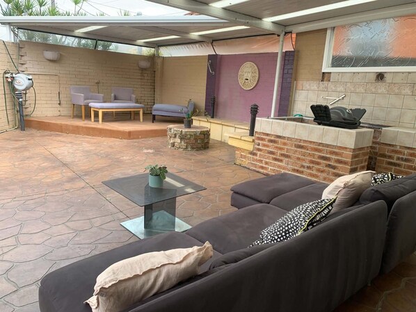 Een buitenterras met een tafel met glazen blad, een bank met kussens, een vloer van stenen tegels en een bakstenen muur met een decoratief bord.