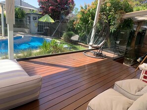 Een houten terras met uitzicht op een zwembad, een hangmat en een glazen constructie met planten.
