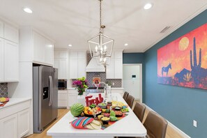 A modern kitchen with white cabinetry, a stainless steel refrigerator, a dining table set with food, and a large painting on the wall.