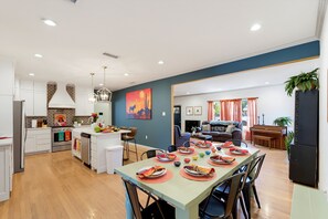 A modern kitchen with white cabinets, a dining area with a table set for six, and a living room with a sofa and a piano.