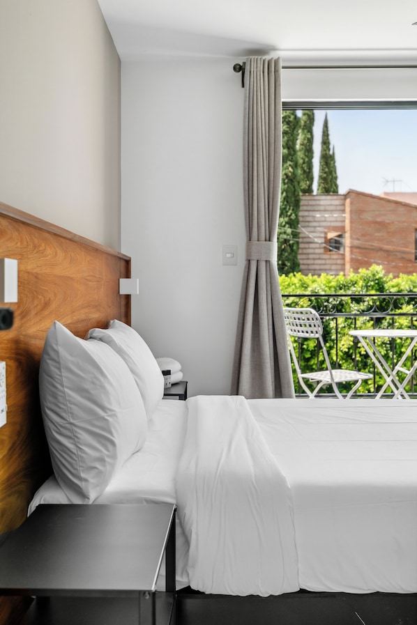 A hotel room with a bed, a nightstand, a wooden headboard, a window with curtains, and a view of greenery outside.