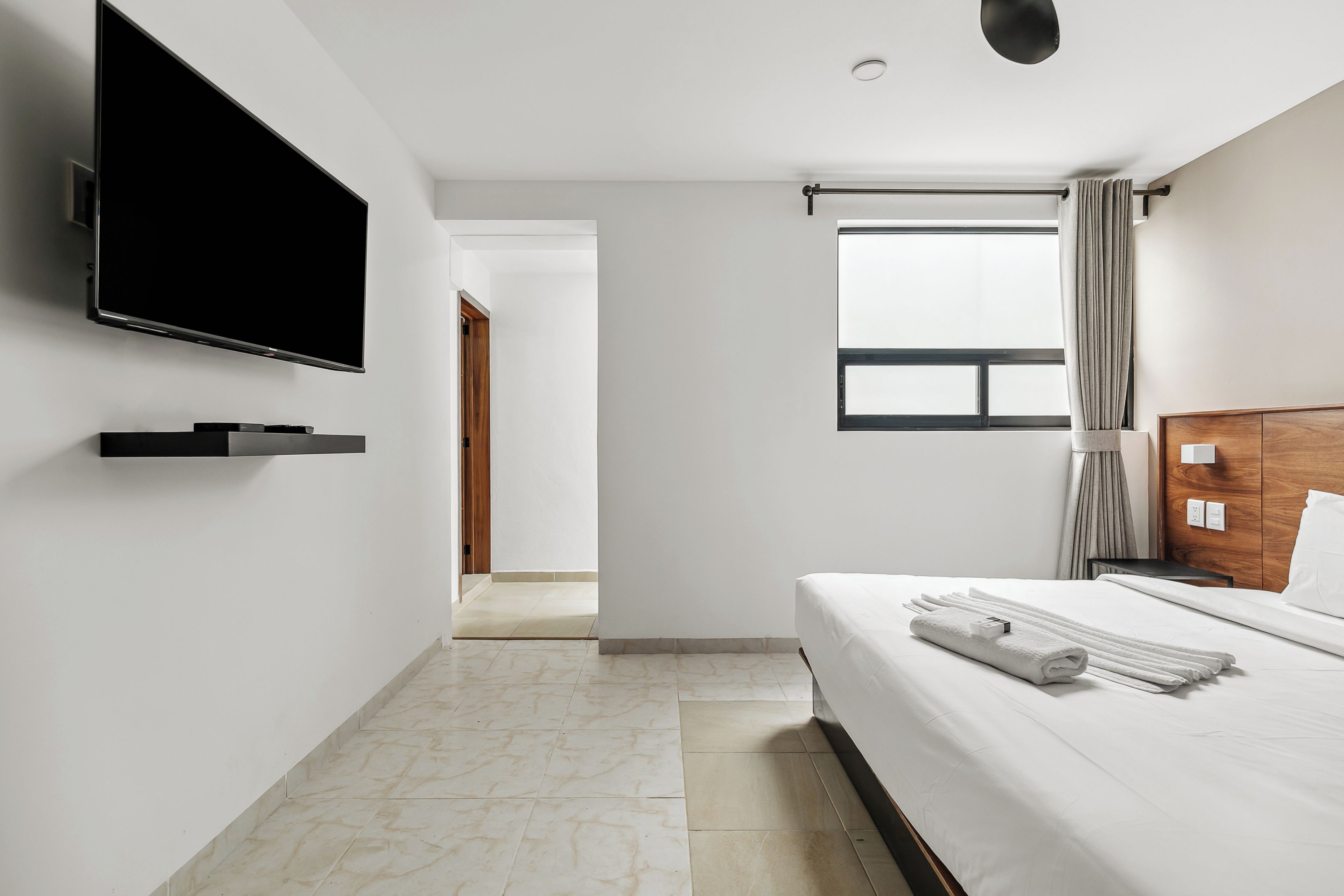Una habitación de hotel moderna con una televisión de pantalla plana, una cama con toallas ordenadas, un cabecero de madera y una ventana con cortinas.