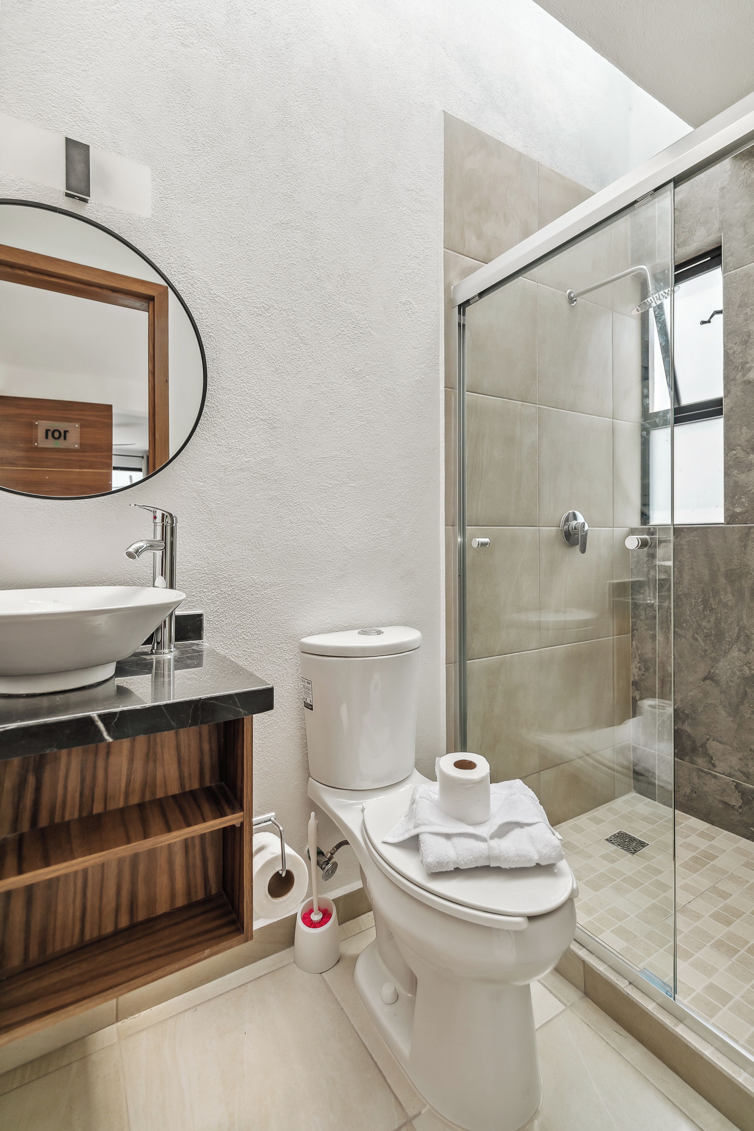 Un baño moderno con un espejo redondo, un lavamanos, un inodoro y una zona de ducha con mampara de vidrio.
