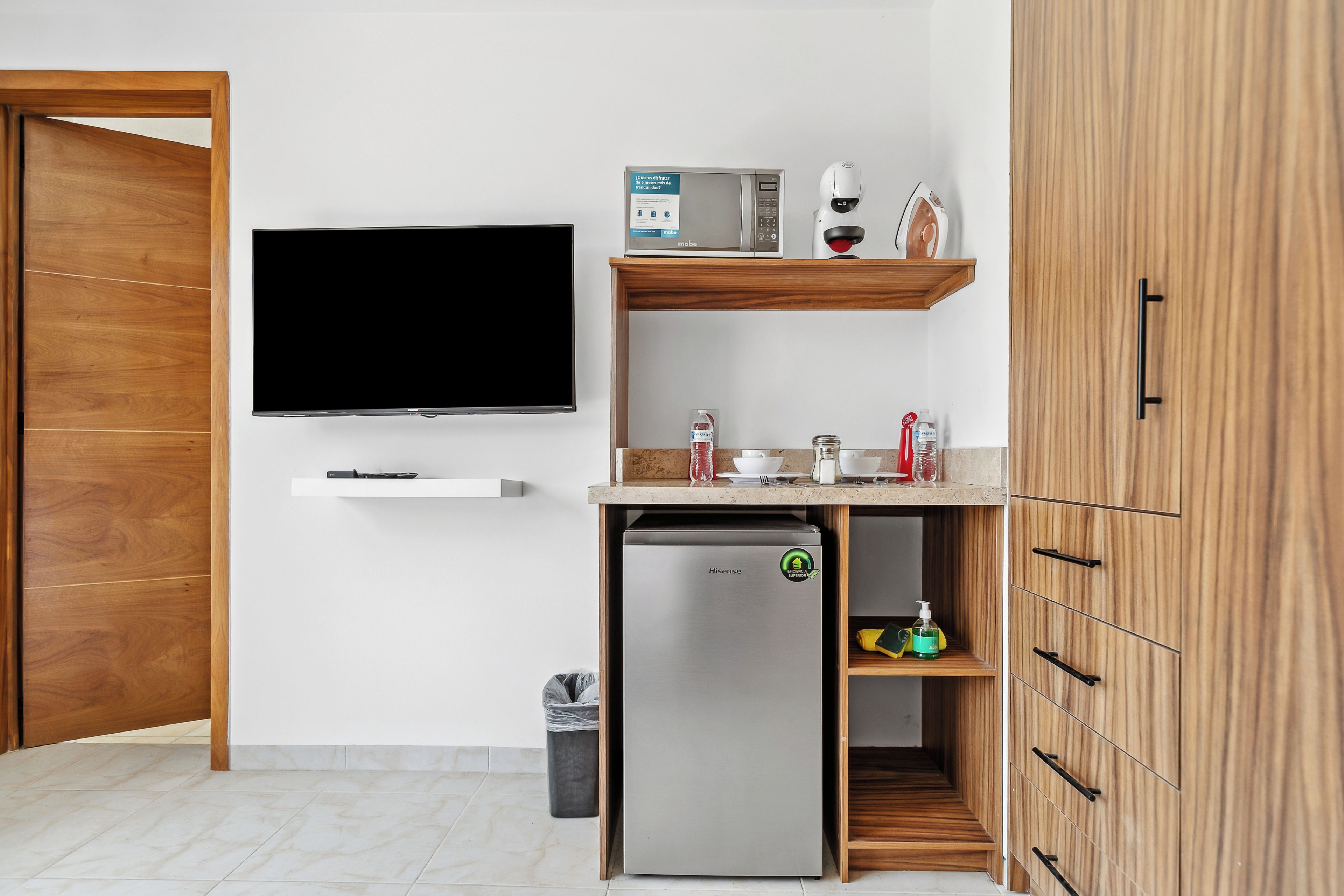 A compact kitchenette with a built-in microwave, a small refrigerator, and a television mounted on the wall.