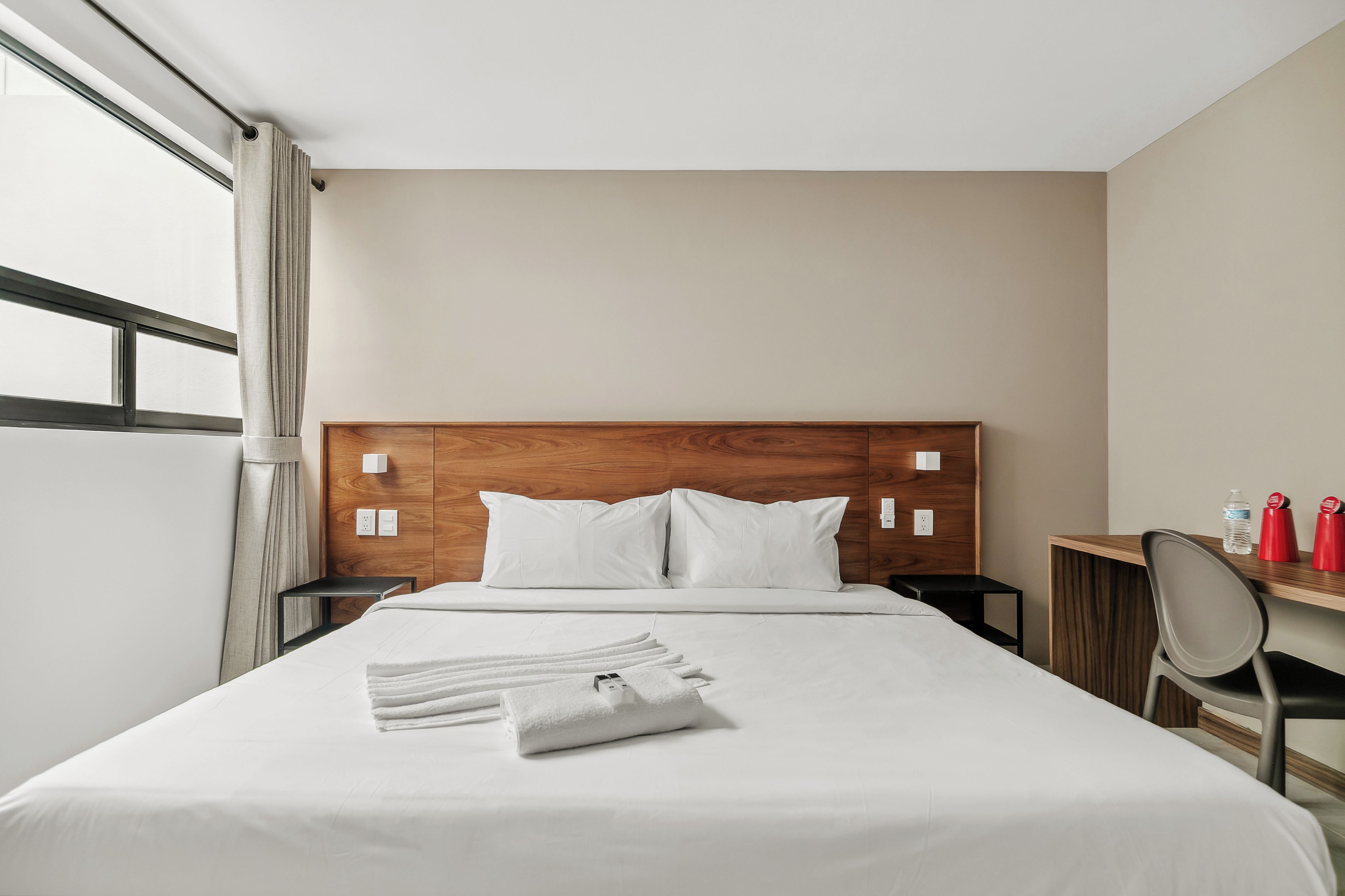 A hotel room with a wooden headboard, a bed with white linens, a small bedside table, a mirror, and a desk with a chair.