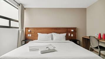 A hotel room with a wooden headboard, a bed with white linens, a small bedside table, a mirror, and a desk with a chair.