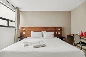 A hotel room with a wooden headboard, a bed with white linens, a small bedside table, a mirror, and a desk with a chair.