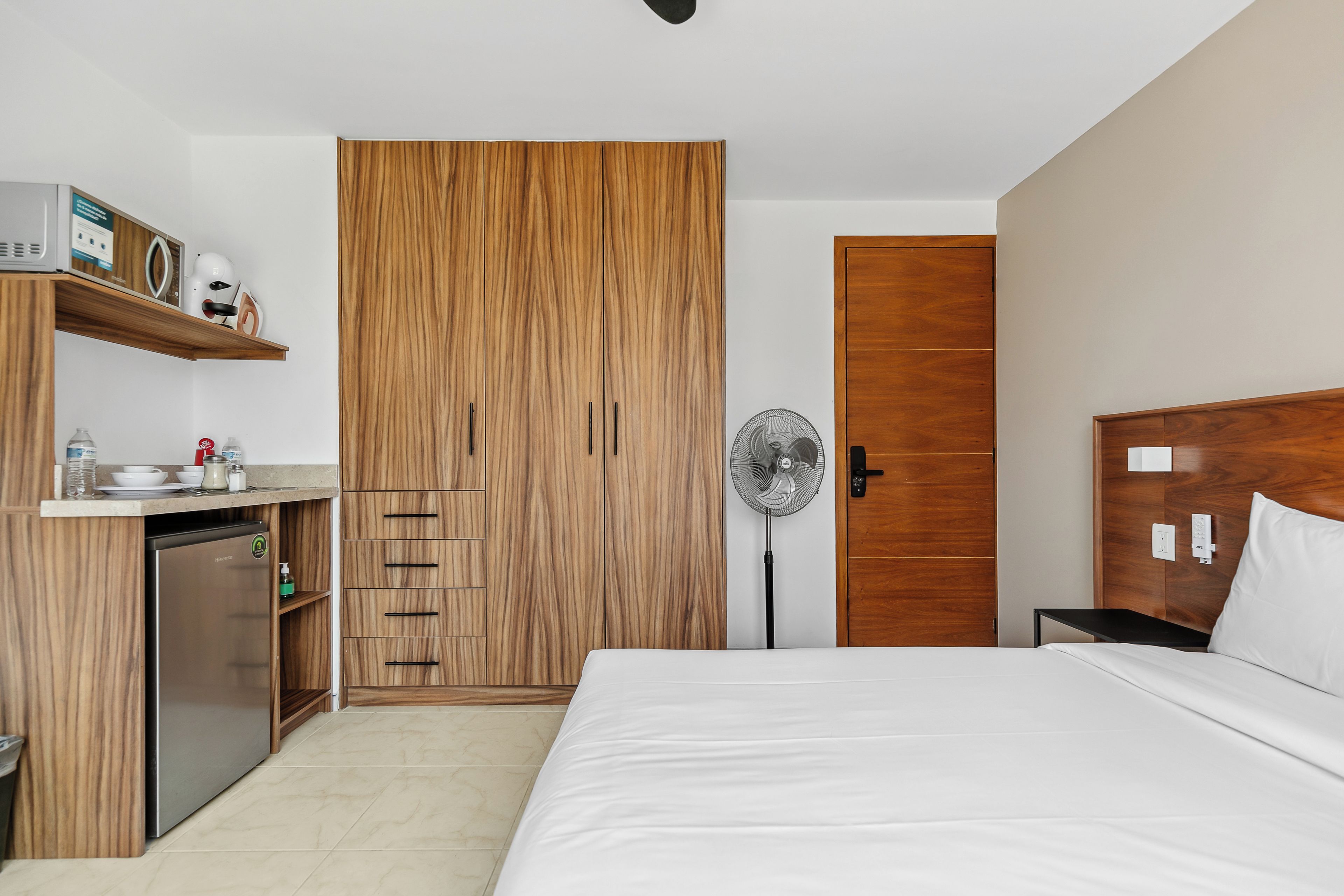 Une chambre d’hôtel avec un lit, une armoire en bois, une petite kitchenette et un ventilateur sur pied.