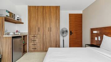 Une chambre d’hôtel avec un lit, une armoire en bois, une petite kitchenette et un ventilateur sur pied.