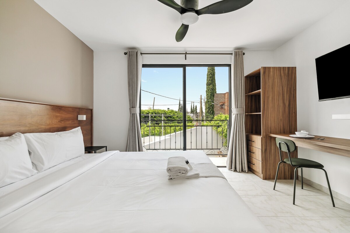 Quarto de hotel com cama, escrivaninha, cadeira, televisão e varanda vista para áreas verdes e edifícios.