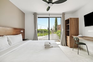 A hotel room with a bed, a desk, a chair, a TV, and a balcony with a view of greenery and buildings.