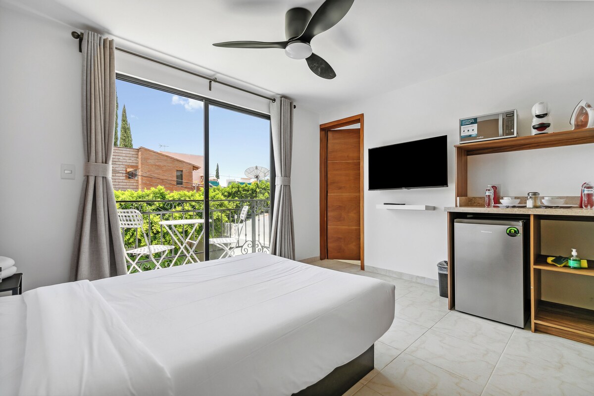 Quarto de hotel com cama, escrivaninha com frigorífico, televisão e varanda com cadeiras.