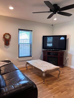 A living room with a leather couch, a television on a stand, a ceiling fan, and a window with blinds. - Charming Sanger Home - King Bed & Loft - Sleeps 6 (Sanger)