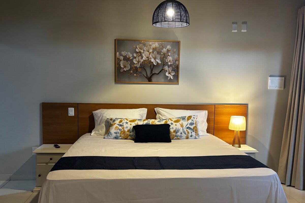 Una habitación de hotel con una cama, mesitas de noche, una lámpara, un cuadro y una ventana con cortinas.