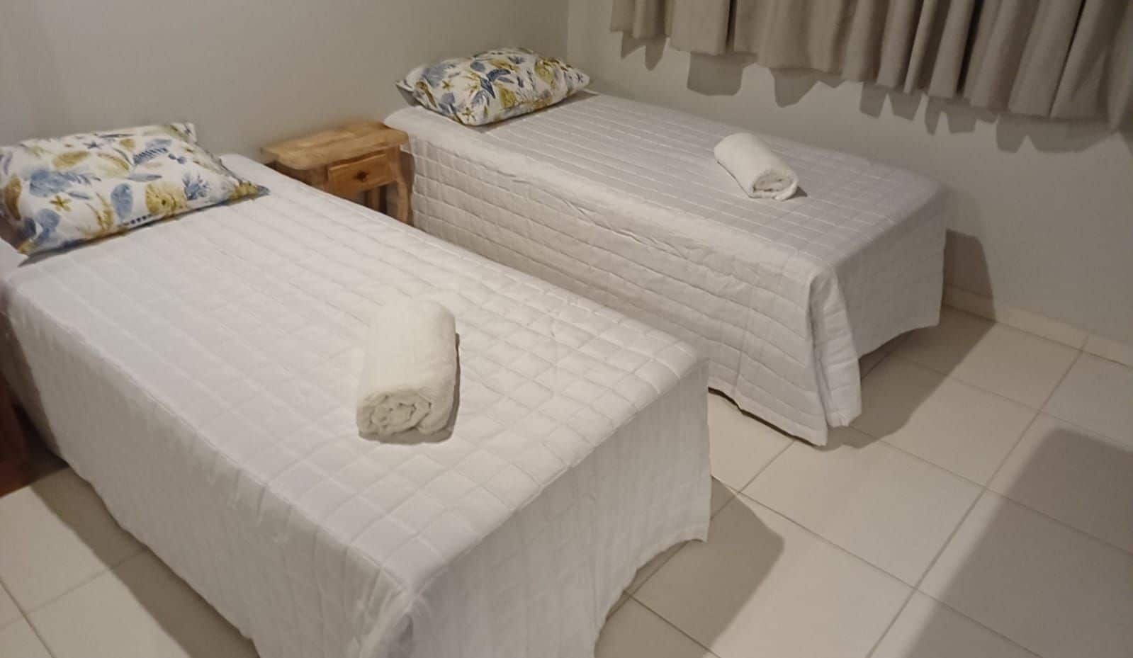 Duas camas de solteiro com roupa de cama branca e uma pequena mesa de cabeceira de madeira entre elas.