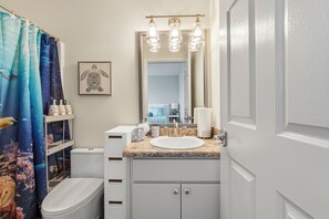 Un baño con inodoro blanco, lavamanos con encimera de mármol y cortina de ducha con motivos marinos.