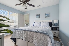 Un dormitorio con cama, mesita de noche, lámpara, ventilador de techo y ventana con persianas.