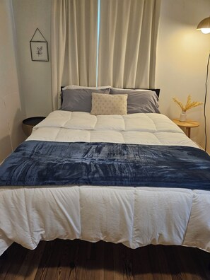 A bed with a white and dark blue quilt, a small bedside table with a vase, and a wall-mounted picture. - Cozy Greenville Home Near Hospitals & Downtown, Pet Friendly (Greenville)