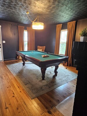 A pool table in a room with wooden flooring, a ceiling fan, and a window with curtains. - Cozy Greenville Home Near Hospitals & Downtown, Pet Friendly (Greenville)