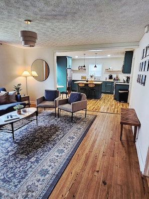 Ein modernes Wohnzimmer mit einer Couch, Sesseln, einem Couchtisch und einem Teppich. Im Hintergrund befindet sich ein Küchenbereich mit einer Bar und Hockern.