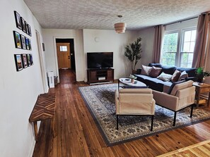 A living room with a sofa, armchairs, a coffee table, a television, and a large window with curtains. - Cozy Greenville Home Near Hospitals & Downtown, Pet Friendly (Greenville)