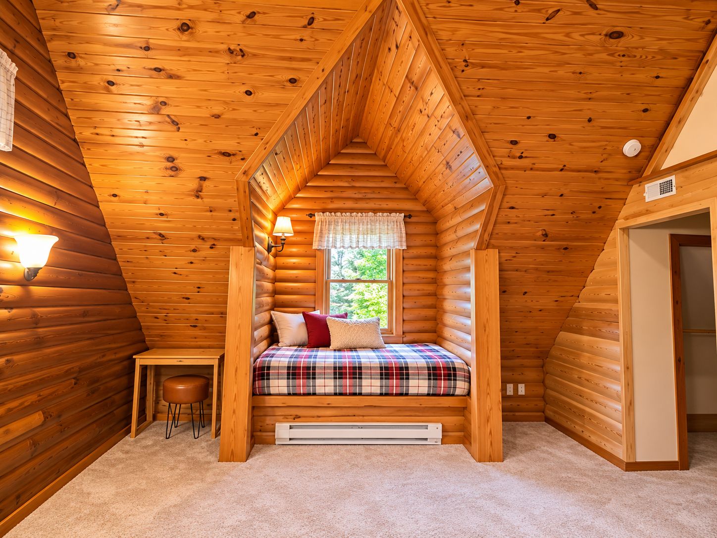Un dormitorio acogedor en el ático, con techo inclinado, cama con ropa de cama escocesa, mesita de noche, lámpara y ventana con vistas.