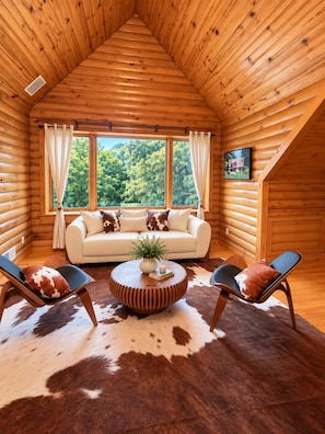 Interior de cabana de tronco com teto alto, um sofá, duas poltronas, uma mesa de centro redonda e um tapete de couro de vaca.