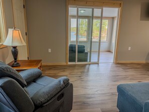A room with a wooden floor, a sofa, a lamp, and a sliding glass door leading to a balcony with a view of trees and a body of water. - Waterfront Group Retreat | 2 Cabins | Sleeps 24 (Hammond)