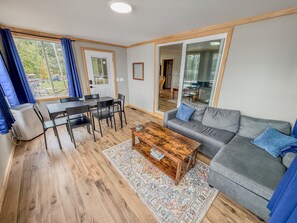 A modern living room with a grey sofa, wooden coffee table, and a dining area with a table and chairs. - Waterfront Group Retreat | 2 Cabins | Sleeps 24 (Hammond)