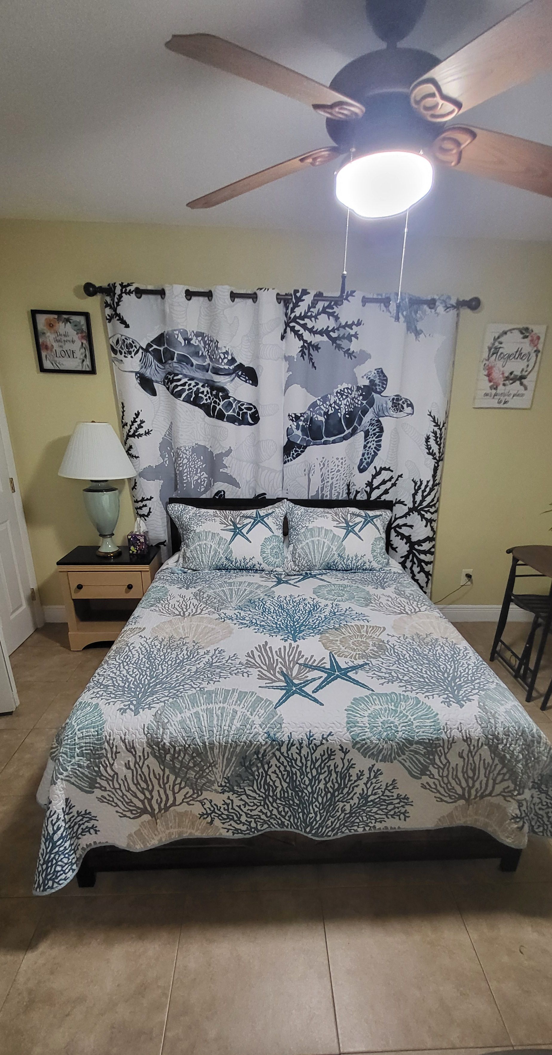 A bed with a marine-themed bedspread, a nightstand with a lamp, and a ceiling fan.