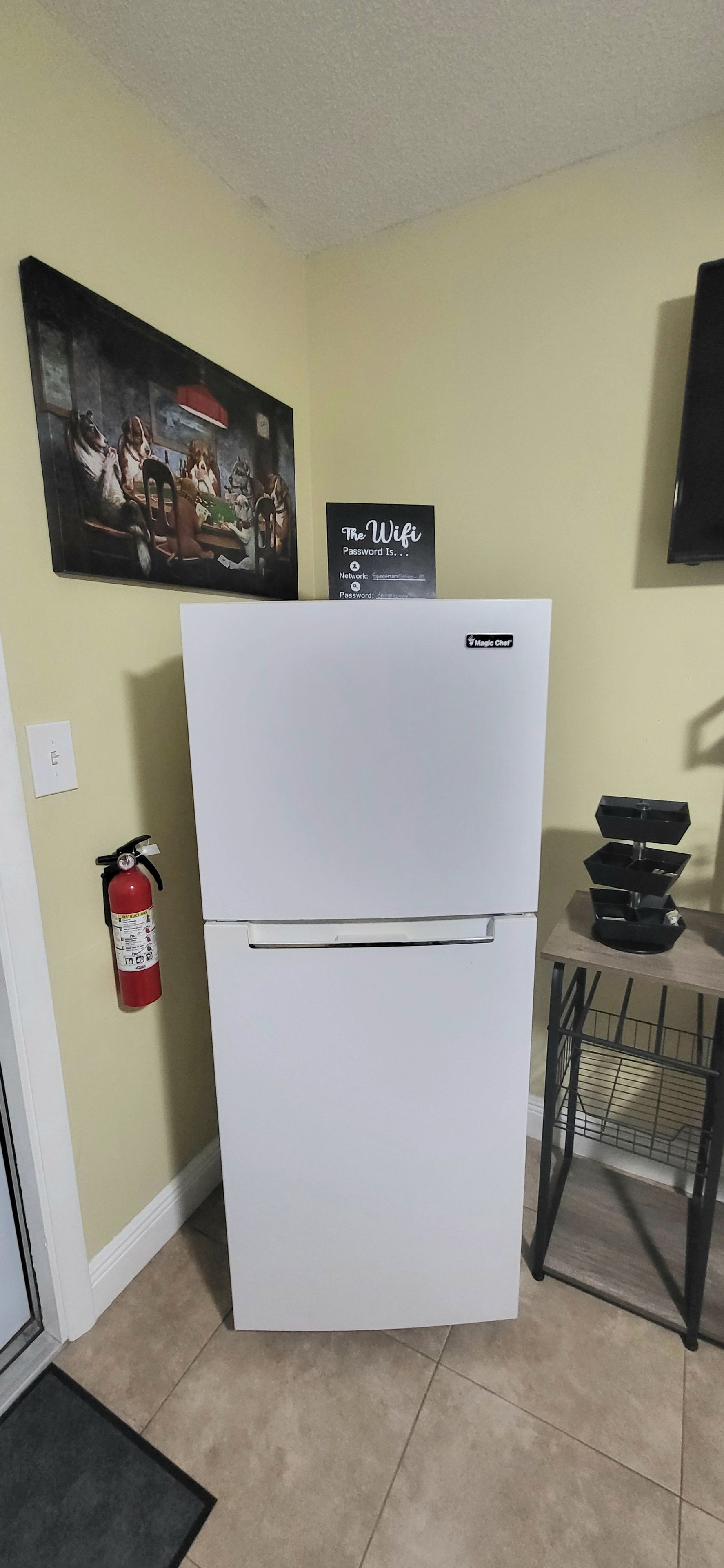 A kitchen with a white refrigerator, a fire extinguisher, a wall-mounted sign, and a small table with a black tray.