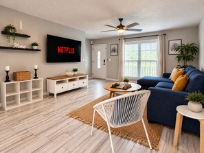 A modern living room with a flat-screen TV, a ceiling fan, a sofa, a coffee table, and a white chair. - 5-bedroom Getaway w/ Private Pool, Arcade, Pool Table & More (Gastonia)