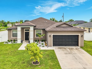 A residential house with a garage, a front door, and a small front yard with a tree. - Coastal Vibes and Pool Days- Cape Coral-5BR/3BA/ Sleep 12 (Cape Coral)
