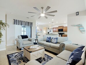 Ein modernes Wohnzimmer mit Deckenventilator, Essbereich und einer Küche im Hintergrund.