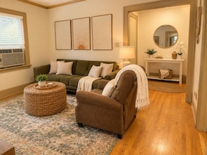 Un salon avec un canapé vert, un fauteuil marron, un pouf rond en osier et un tapis à motifs.