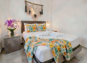 A modern bedroom with a bed, floral pillows, a macrame wall hanging, and a vase with flowers. - Queen Bed, Rooftop Pool, Quiet Street · Sleeps 4 · Beach & 5th Ave 10 Min Walk (Playa del Carmen)