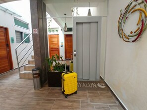 Entrada de hotel com uma mala amarela, uma lixeira e escadas que levam aos quartos.