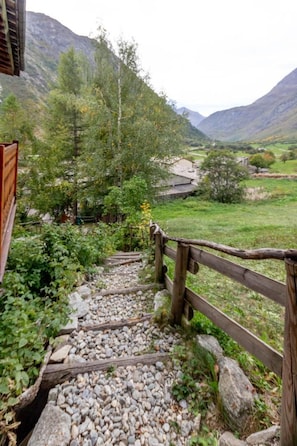 A wooden pathway leading through a mountainous area with a view of a valley and distant buildings. - Les Abeillos - Le Marmotton (BONNEVAL SUR ARC)