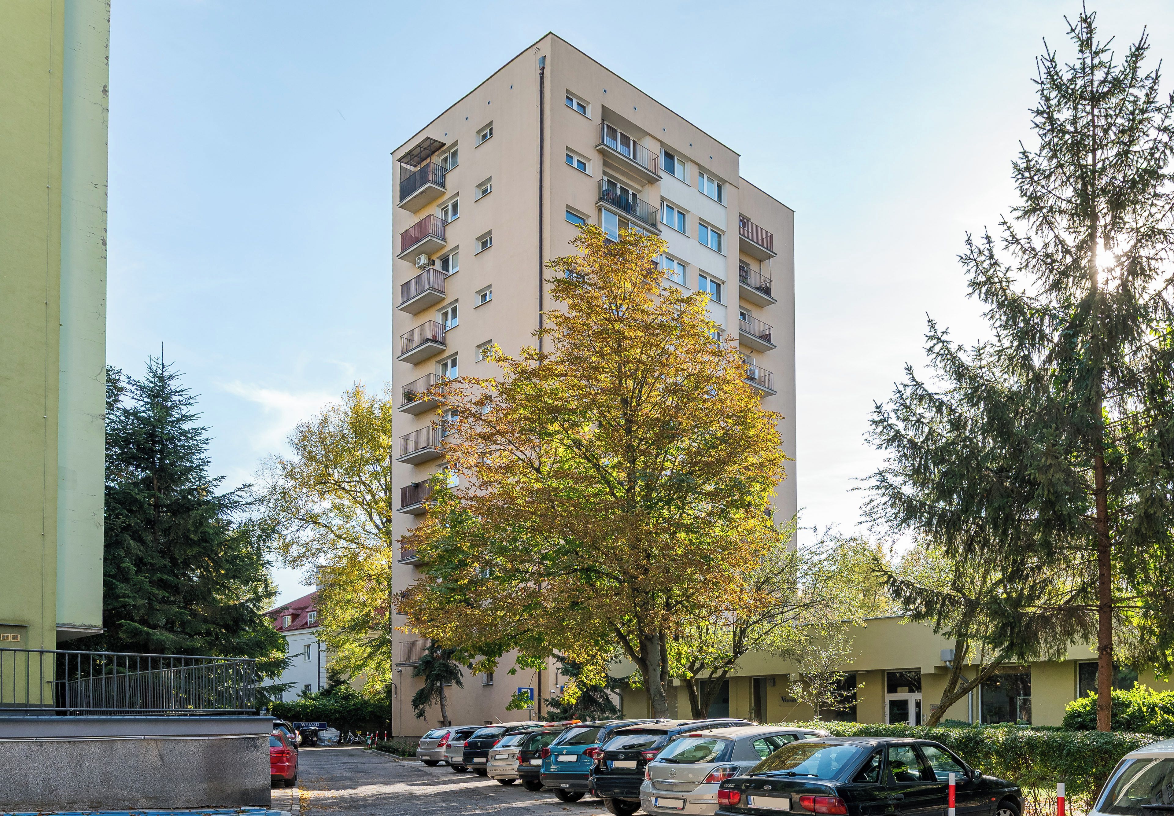 Un immeuble résidentiel à plusieurs étages, doté de balcons, d’un espace de stationnement avec des voitures et entouré d’arbres.
