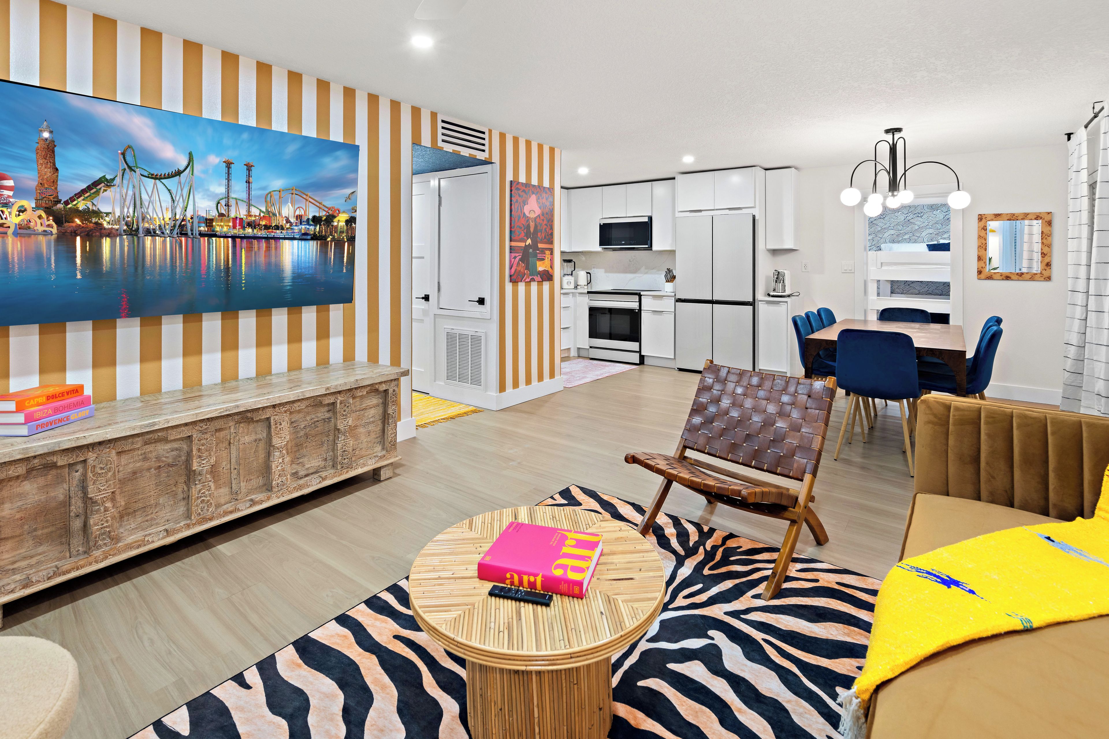 A modern living room with a large TV, a dining area with blue chairs, and a kitchen with white cabinets.