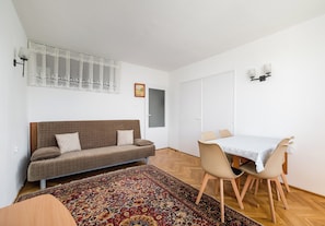Ein Zimmer mit einem braunen Sofa, einem Esstisch mit Stühlen, einem gemusterten Teppich und einem Fenster mit Vorhang.