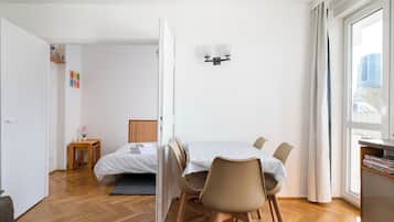 Une chambre d’hôtel moderne, équipée d’un lit, d’une table à manger avec des chaises, d’une petite table de chevet agrémentée d’une plante et d’une fenêtre avec des rideaux.