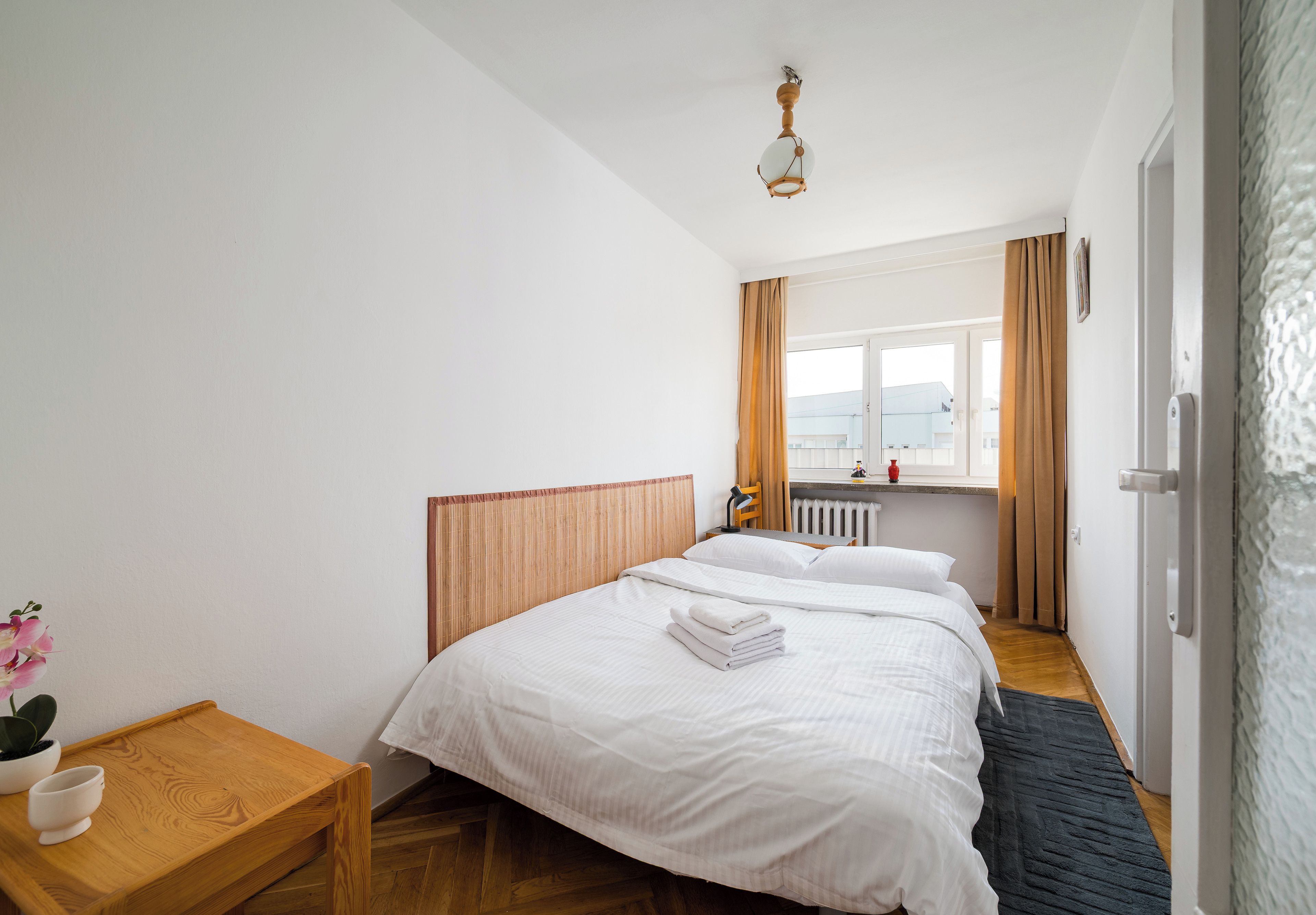 A hotel room with a bed, bedside table, lamp, window with curtains, radiator, and a view of the outside.