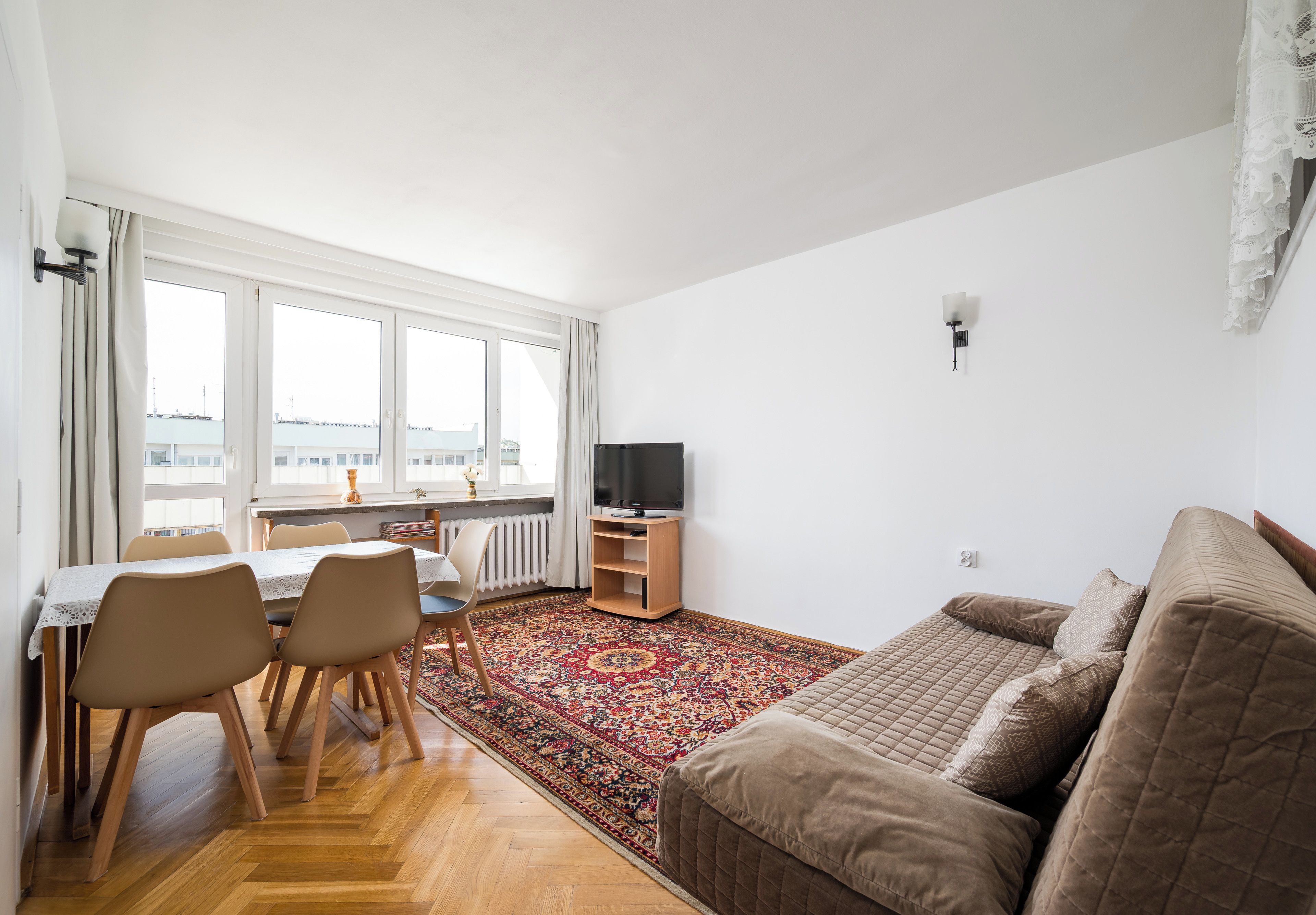 A modern living room with a sofa, a dining table with chairs, a TV stand, and a large window with curtains.