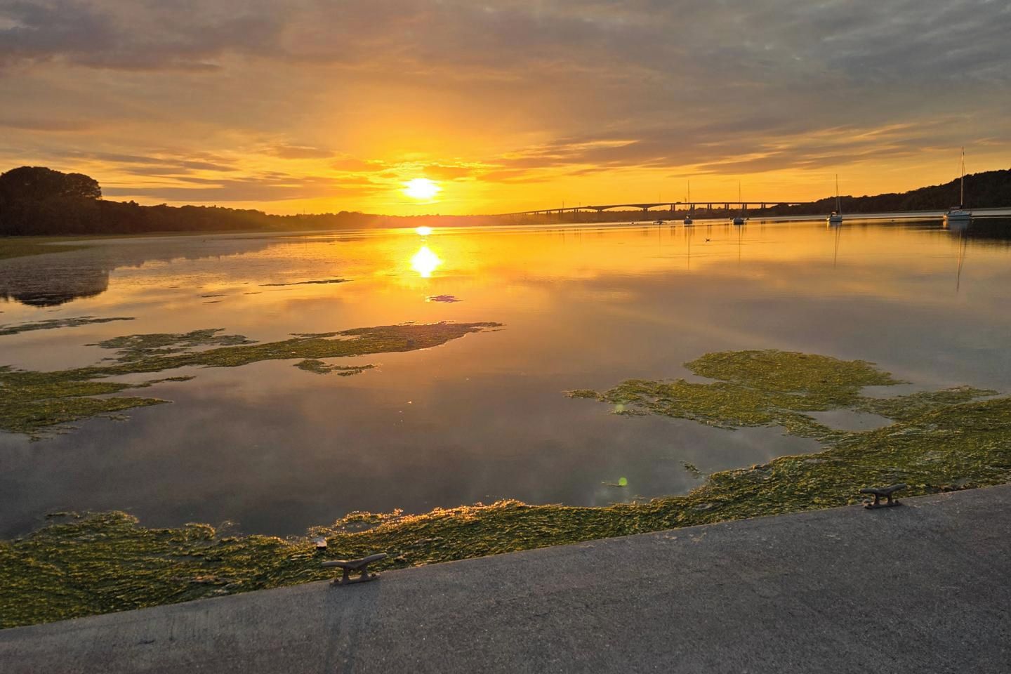 Une étendue d’eau calme où se reflète un coucher de soleil, avec un pont au loin et une promenade pavée qui longe le bord.