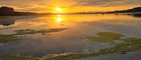 A calm body of water reflecting a sunset, with a bridge in the distance and a paved walkway along the edge.
