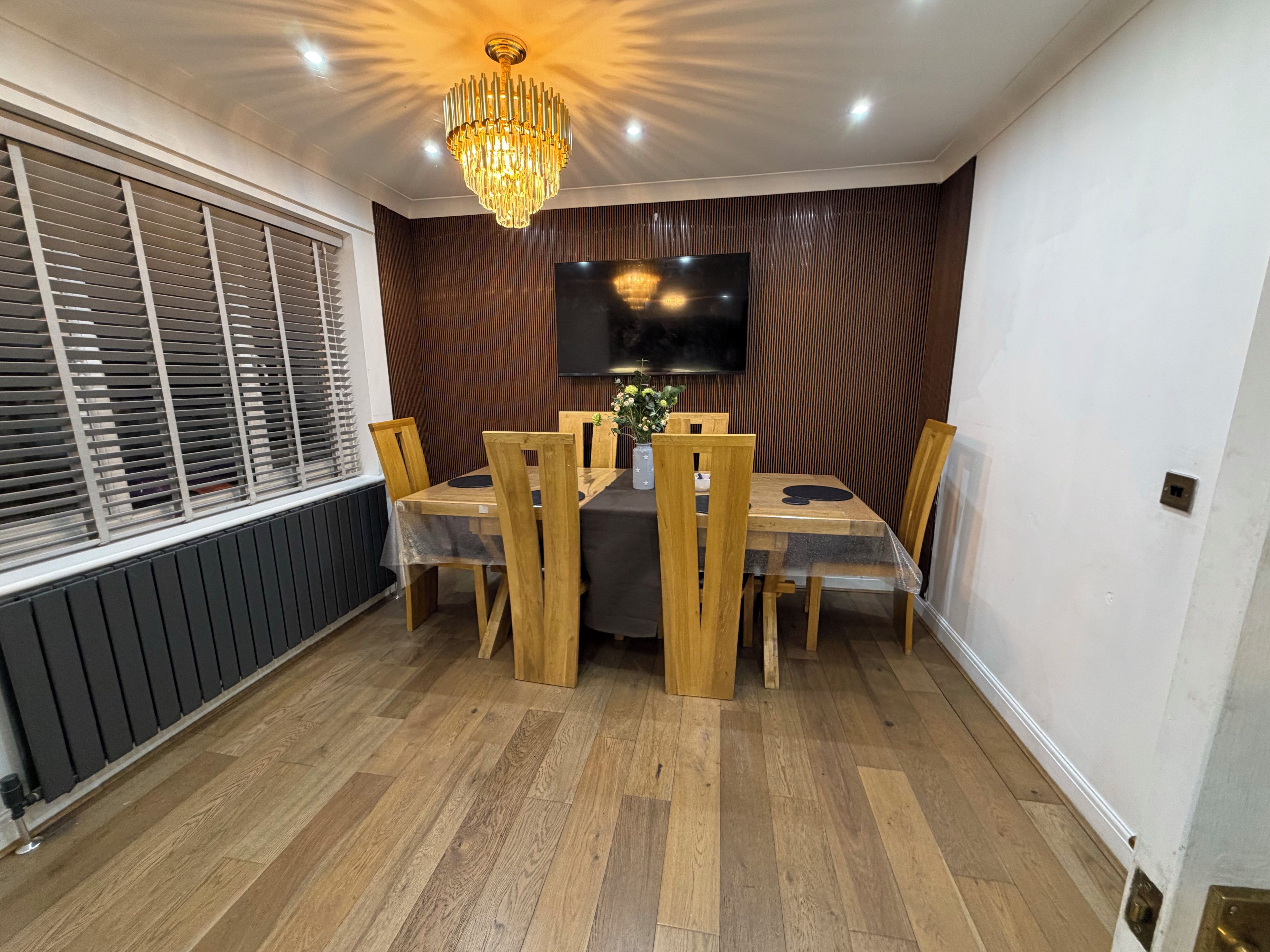 A dining area with a wooden table, chairs, a chandelier, and a television on the wall.