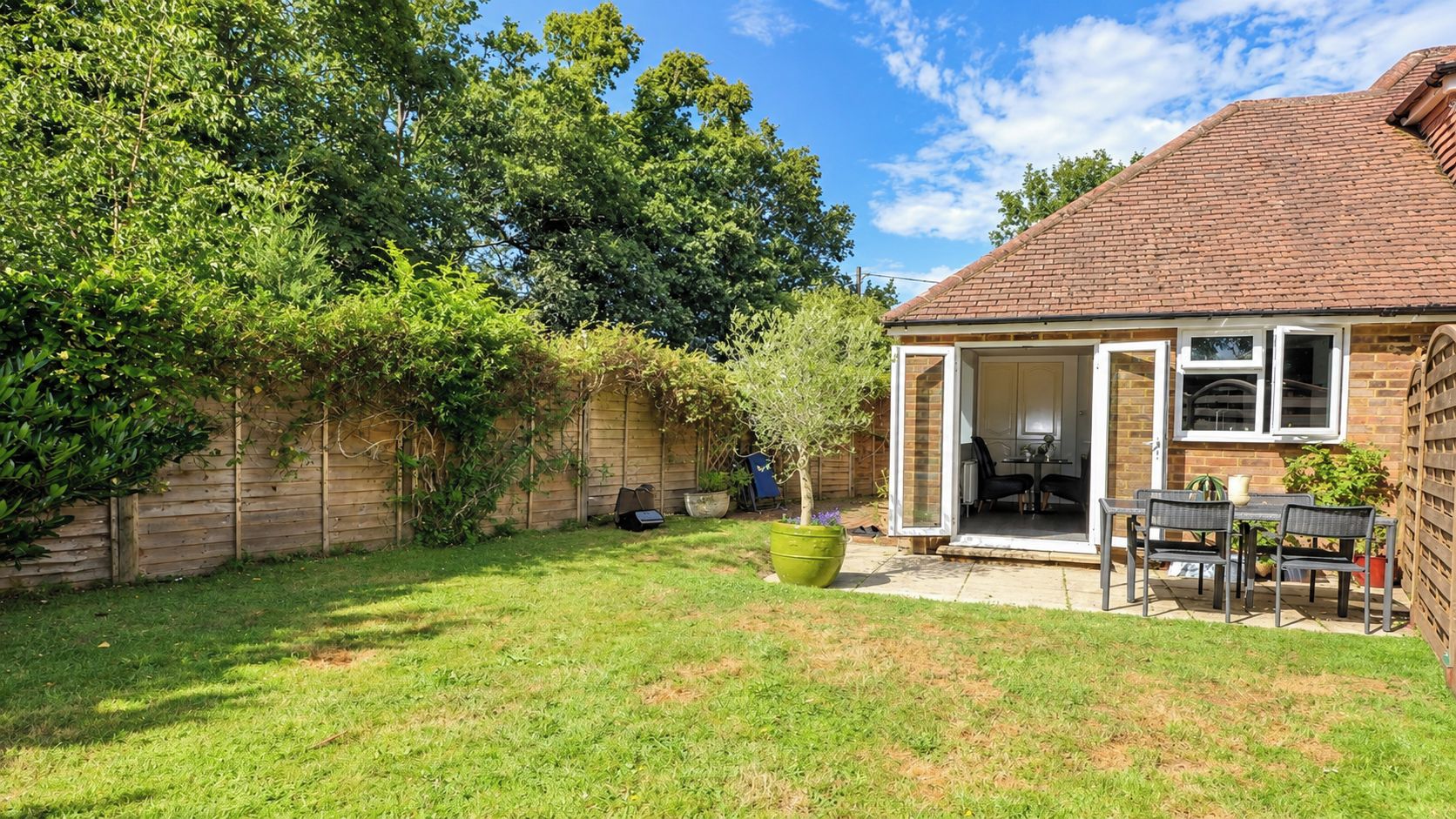 Un jardin avec une terrasse, une table et des chaises, un jardinier en forme de bac et une clôture en bois.