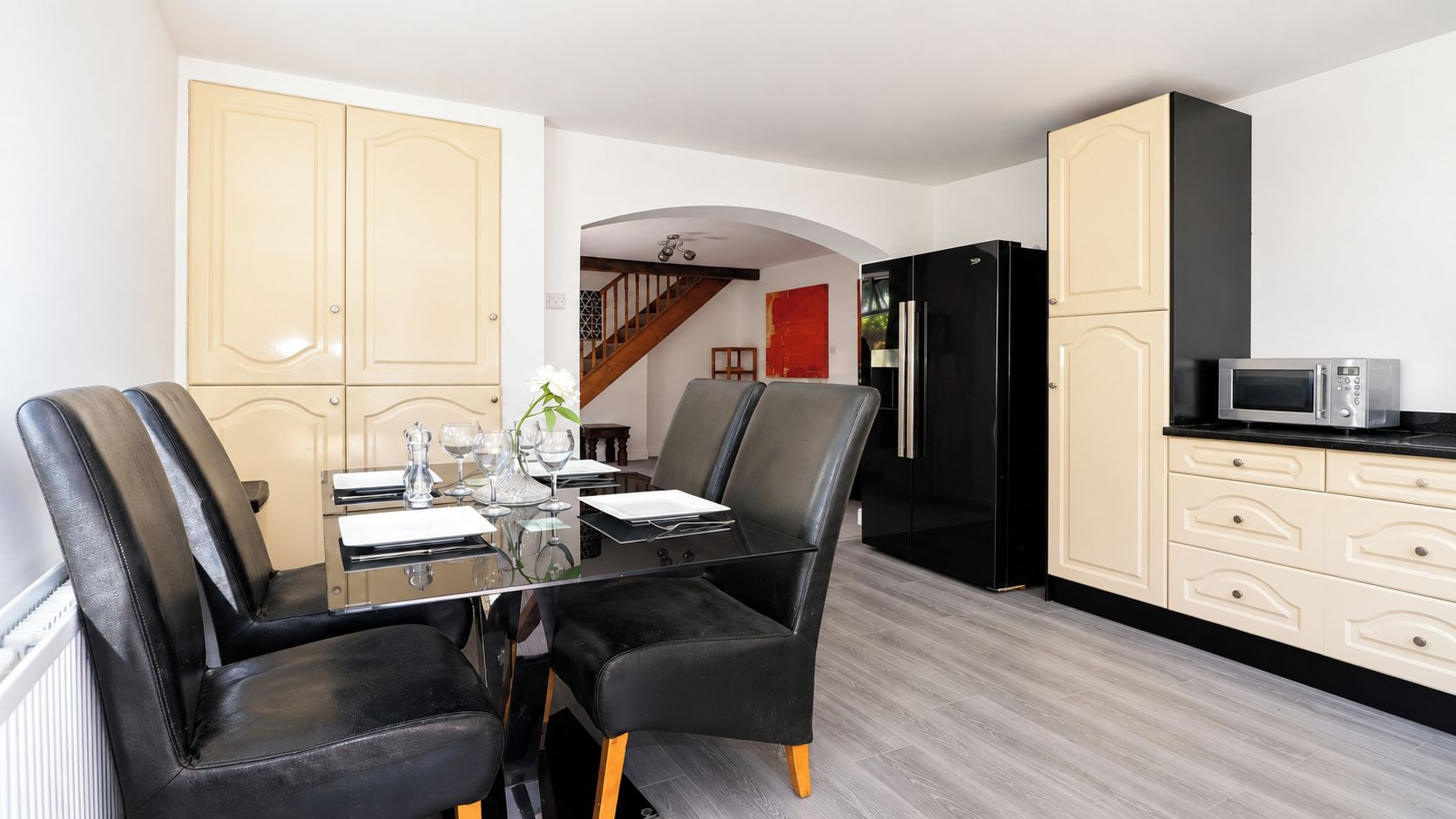 A dining area with a glass table and black chairs, a kitchen with a black refrigerator and white cabinets, and a wooden staircase in the background.